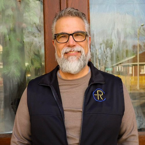 A smiling older man with gray hair, a full beard, and glasses, wearing a black vest with a logo, standing outdoors near a wooden structure.