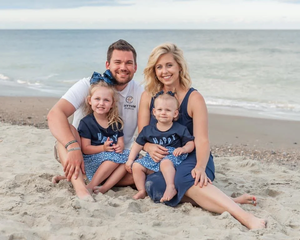 Family sitting on the beach, smiling, with the ocean in the background. The father and mother are sitting with their two young daughters, all wearing matching blue outfits.