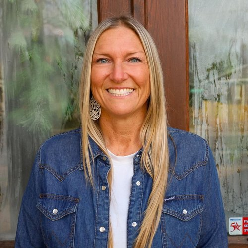 A smiling woman with long blonde hair, wearing a denim jacket and a white shirt, standing in front of a window with a wooden frame.