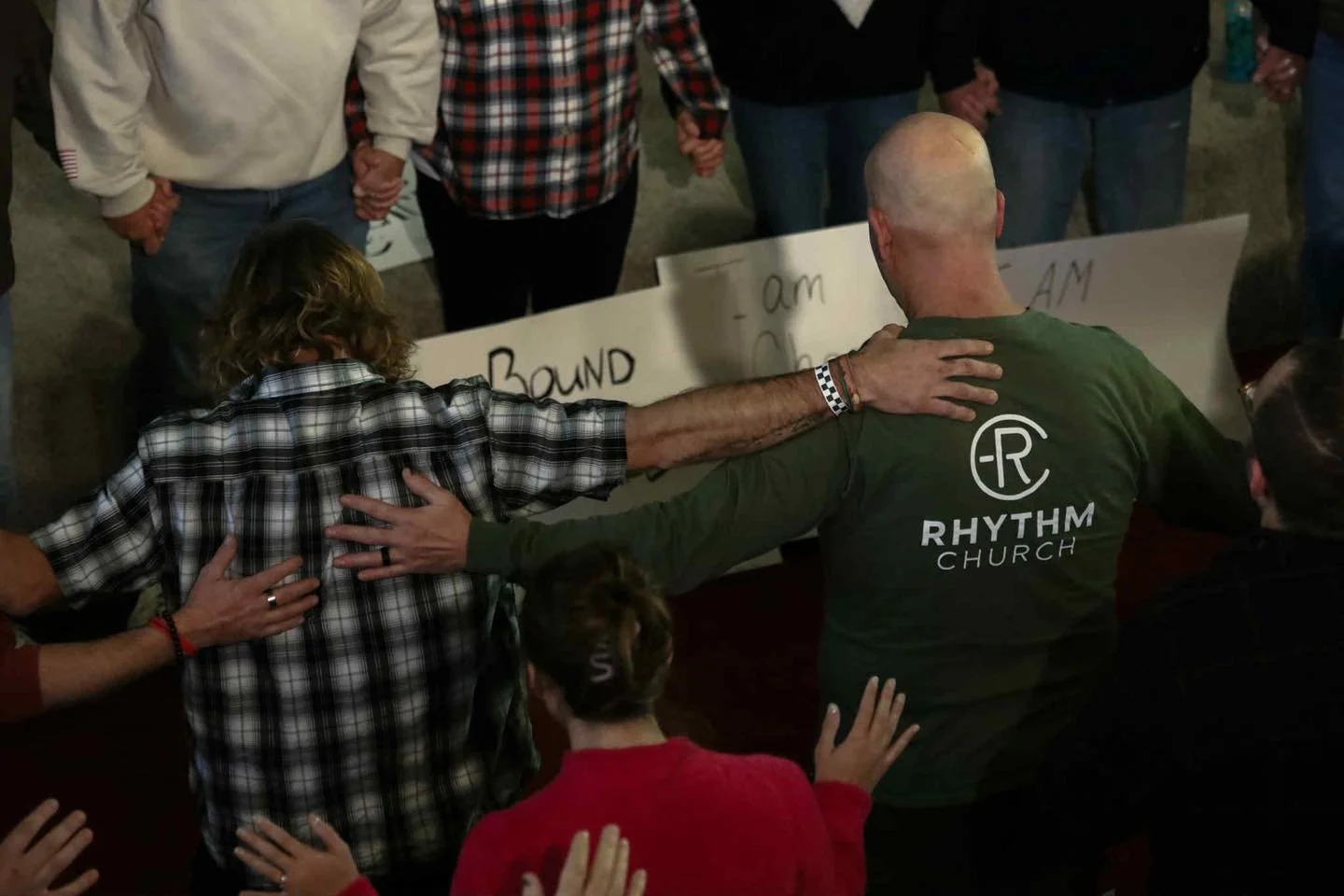 A group of people praying together with their hands on each other's shoulders, surrounding a man in a green Rhythms Church shirt who is kneeling and praying in the center.