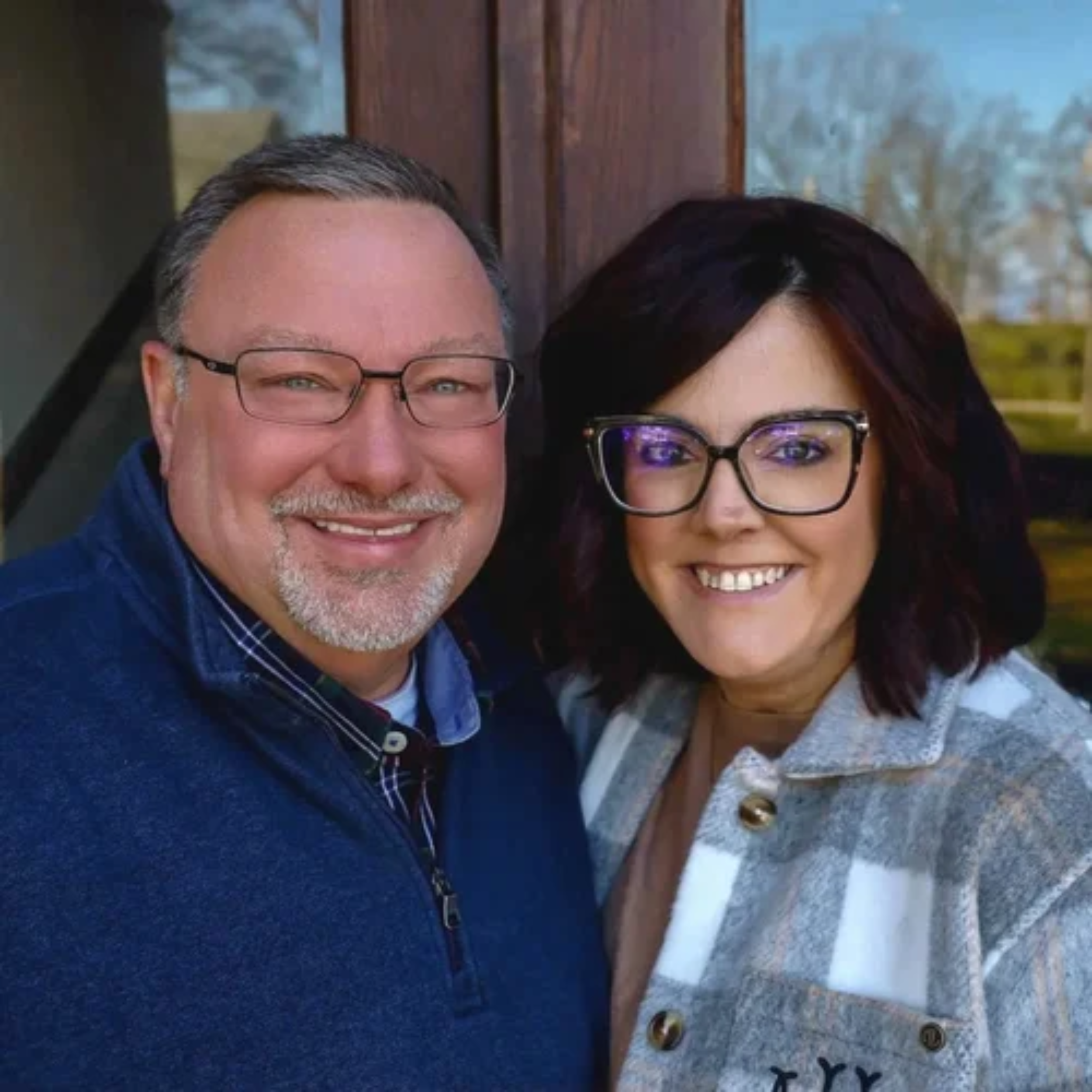 A close-up of a couple smiling, both wearing glasses, with a background featuring trees and a wooden structure, suggesting outdoor or park setting.