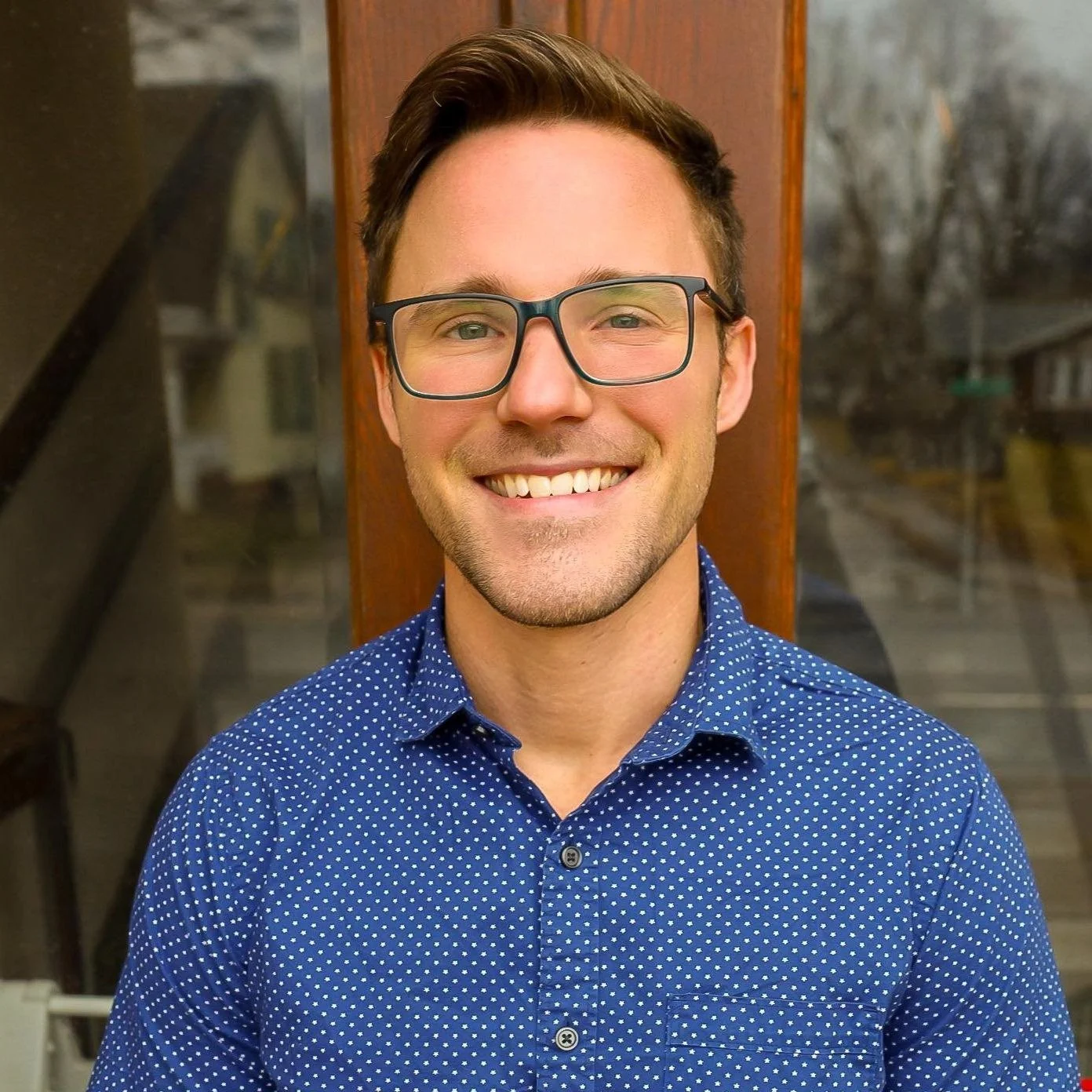 A young man with short brown hair, glasses, and a light beard, smiling, wearing a blue dotted shirt, standing in front of a wooden background with windows showing a cloudy outdoor scene.