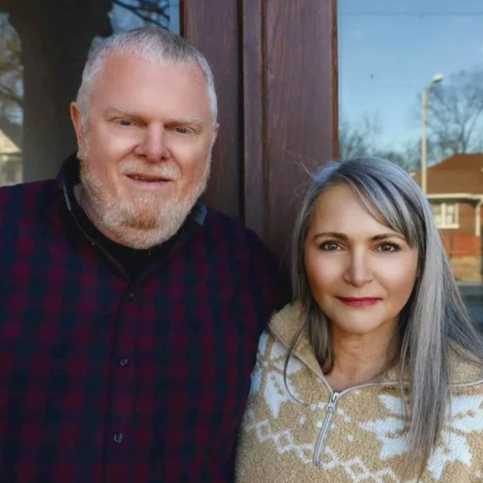 Close-up of a smiling man with short gray hair and a woman with long gray hair, partially visible, standing outdoors with trees and a wooden fence in the background.