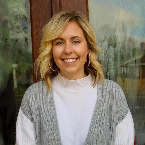 A young woman with blonde hair smiling, wearing a white turtleneck and a gray cardigan, standing in front of a window with greenery outside.