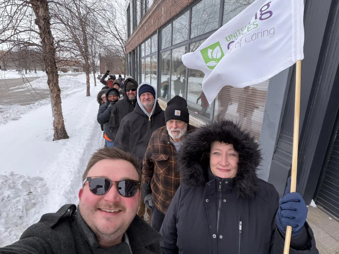Line of people standing outdoors in snowy weather, holding a white flag with green and purple logo and text, in front of a building with large windows.