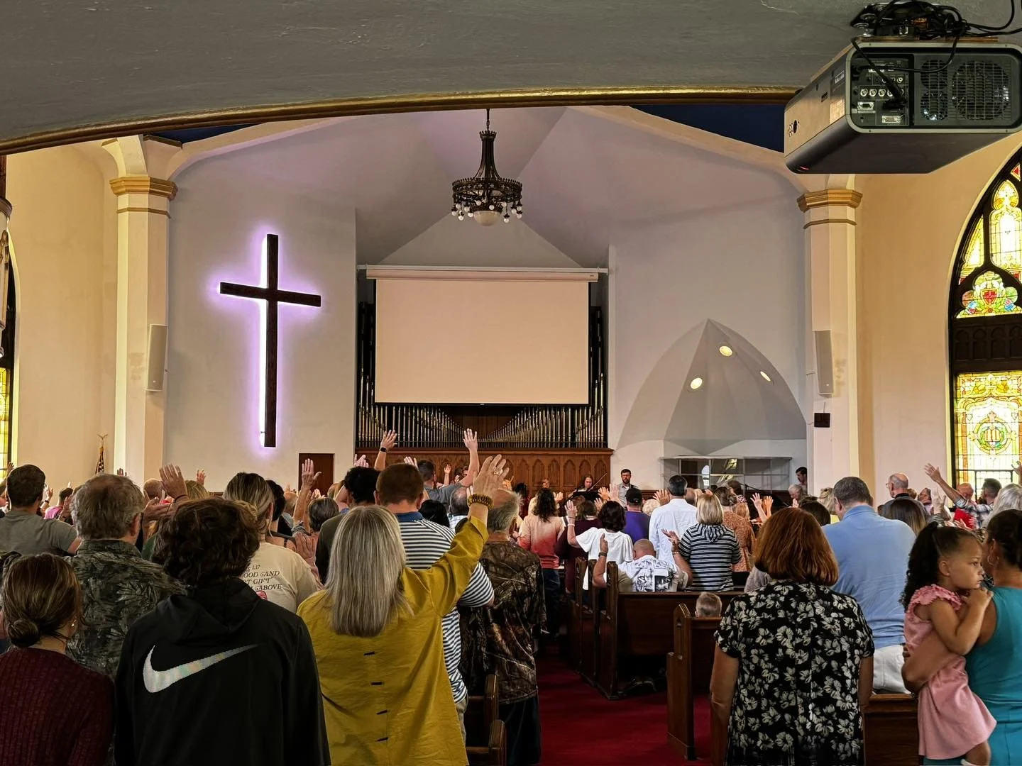 A diverse congregation inside a church during a service, with many people raising their hands. The church features stained glass windows, a prominent lit cross on the wall, and a large projection screen behind the altar.