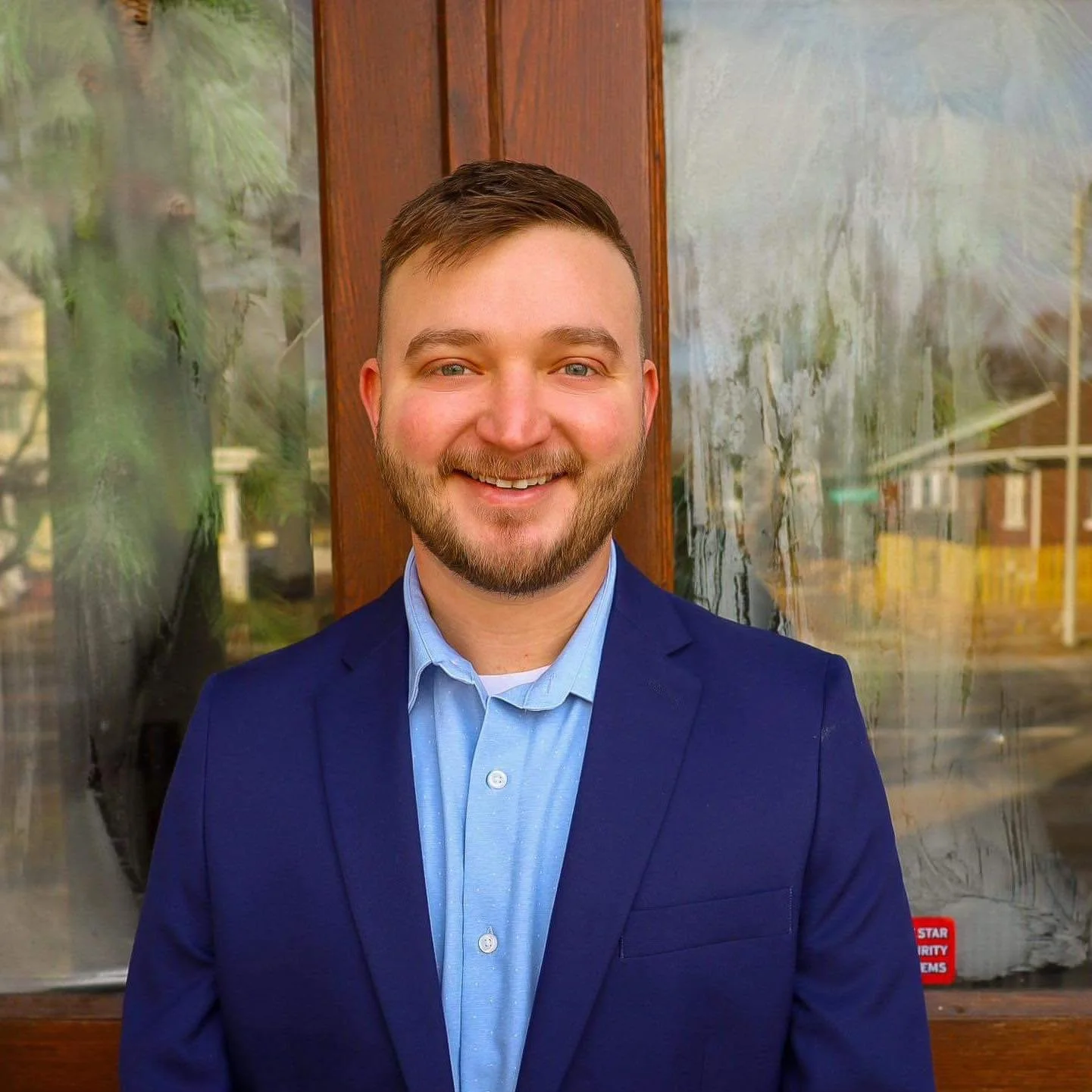 A smiling man with a beard and short brown hair, dressed in a blue suit jacket and light blue shirt, standing in front of a glass window with reflections of trees and buildings.