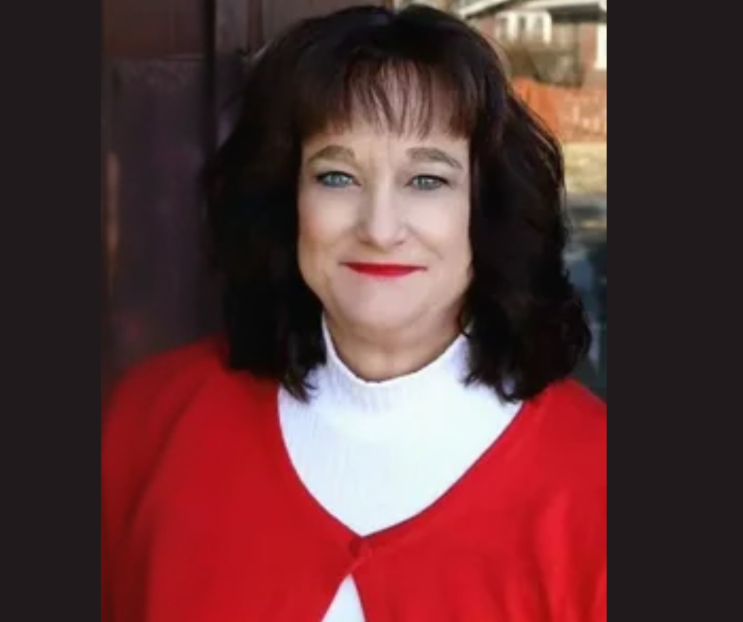 A middle-aged woman with short dark hair and blue eyes, wearing a red top with a white collar, standing outdoors near a brick wall with a blurry background.