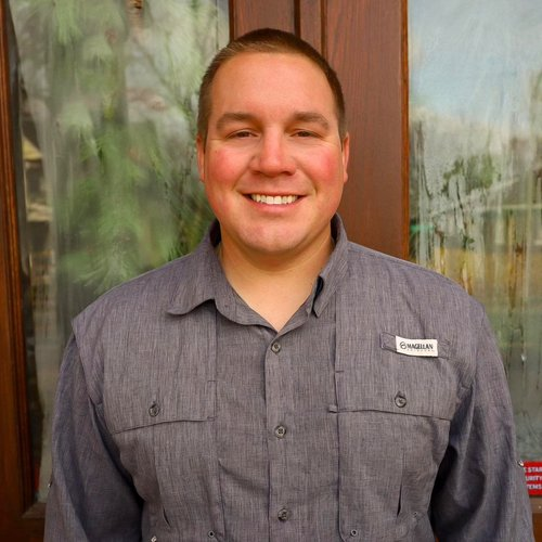 A smiling man wearing a gray collared shirt with a name tag, standing indoors in front of a glass window with a blurred outdoor scene.