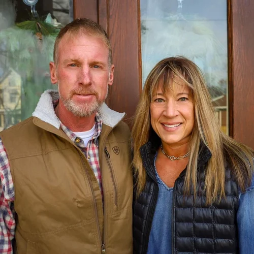 A man and woman standing together outdoors, smiling, in front of a wooden and glass structure.