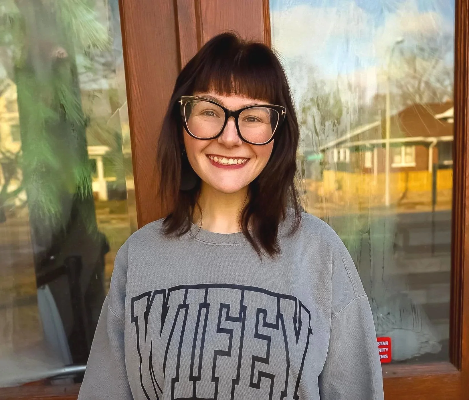 A woman with short dark hair and glasses smiling in front of a glass door with trees and a house visible outside.