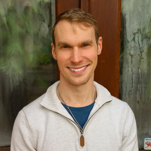 A smiling man with short brown hair and blue eyes, wearing a white zip-up sweater and a blue shirt, standing outdoors near a wooden wall and greenery.