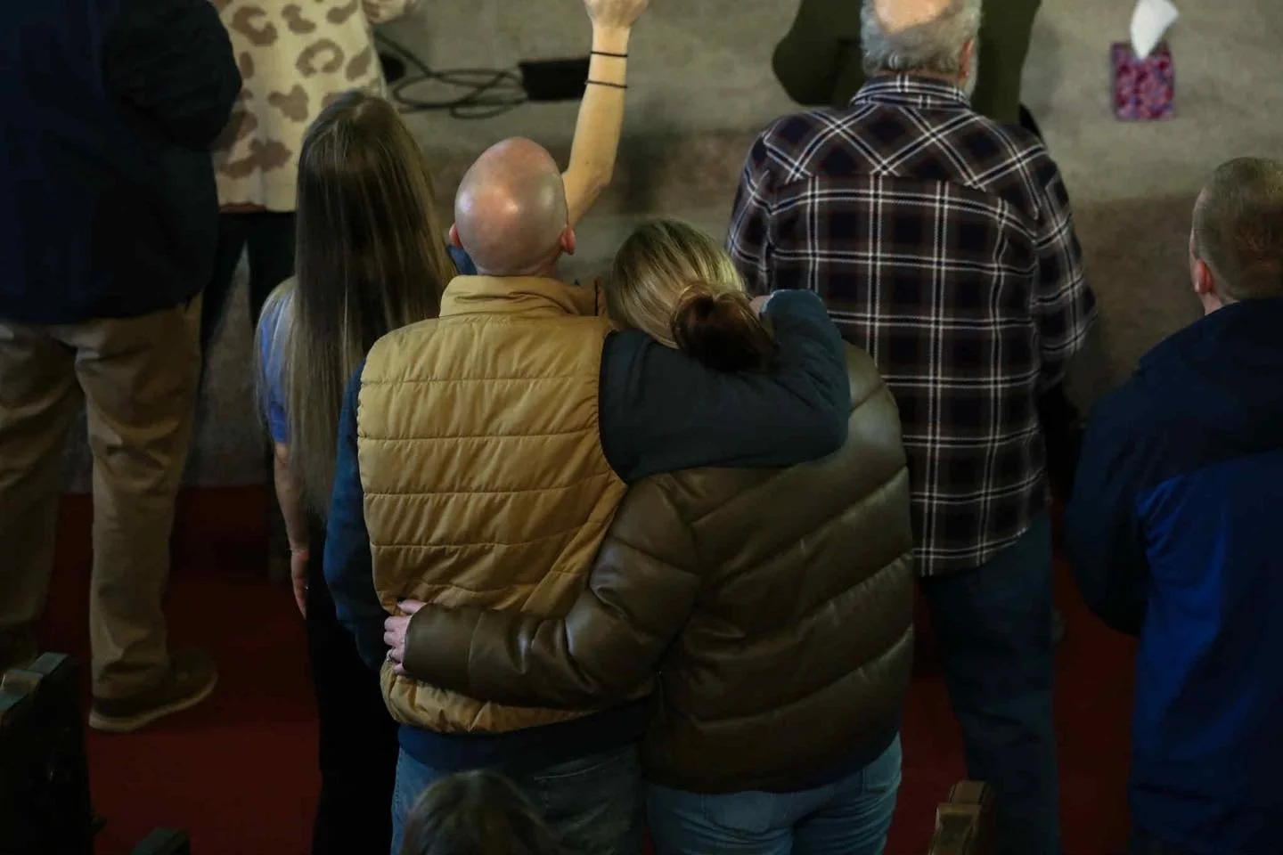 A man and woman sit closely with their arms around each other at an indoor gathering, surrounded by other people.
