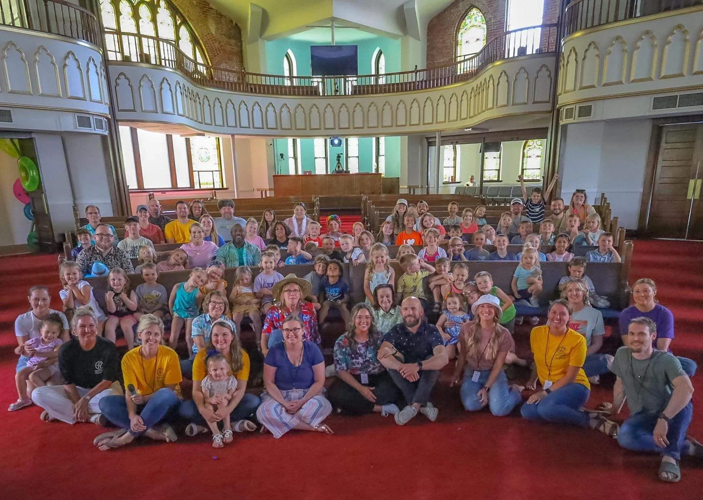 Group photo of children and adults sitting inside a church or hall with stained glass windows, red carpet, and wooden balconies, posing for the camera.