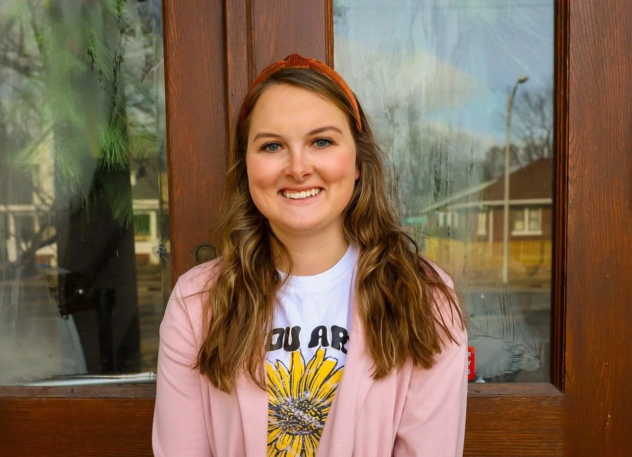 A young woman with long wavy brown hair, blue eyes, and light skin, smiling and wearing a pink jacket and a white T-shirt with a sunflower graphic, standing in front of a wooden and glass door.