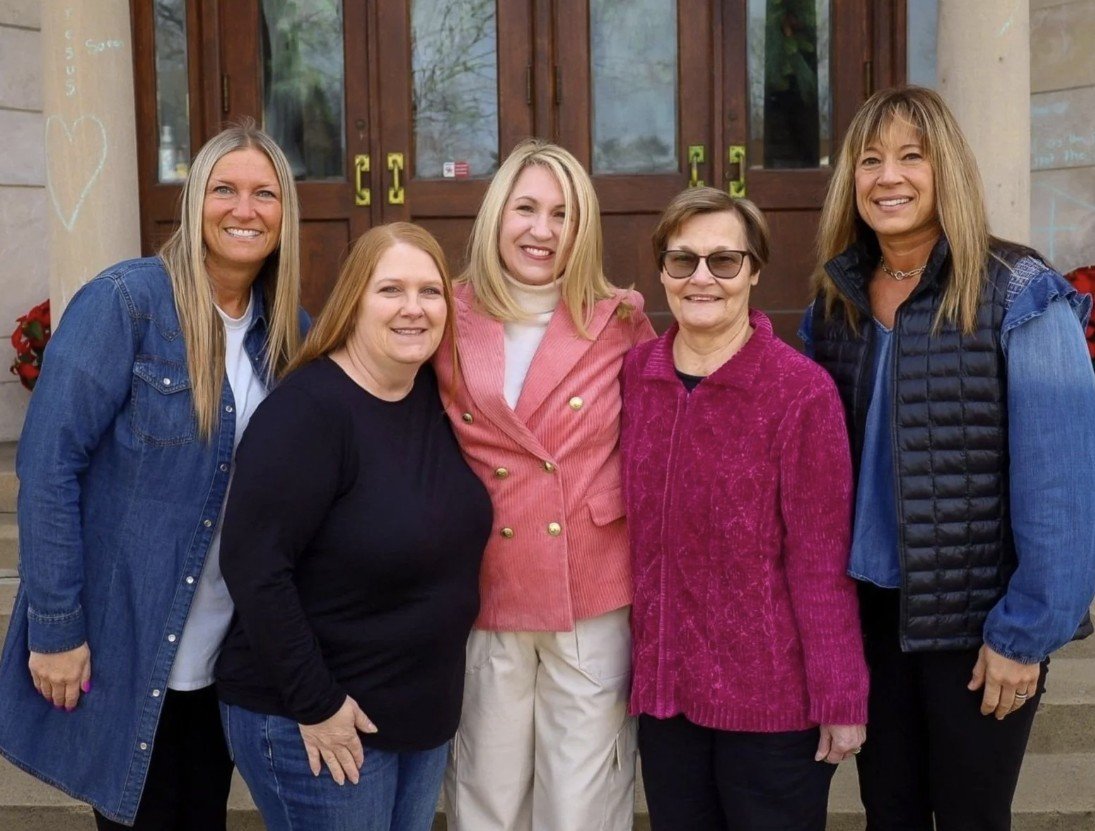 Five women standing together outside, smiling, in front of a wooden door with glass panels.