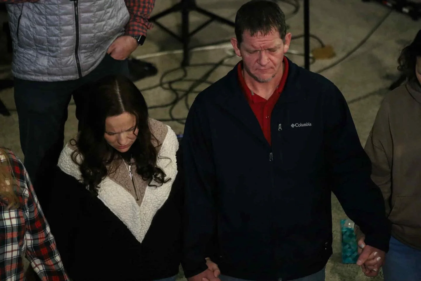A group of people holding hands in a prayer or moment of silence, with a man in a black Columbia jacket and a woman with long dark hair and glasses in a cream and black sweater in the foreground.