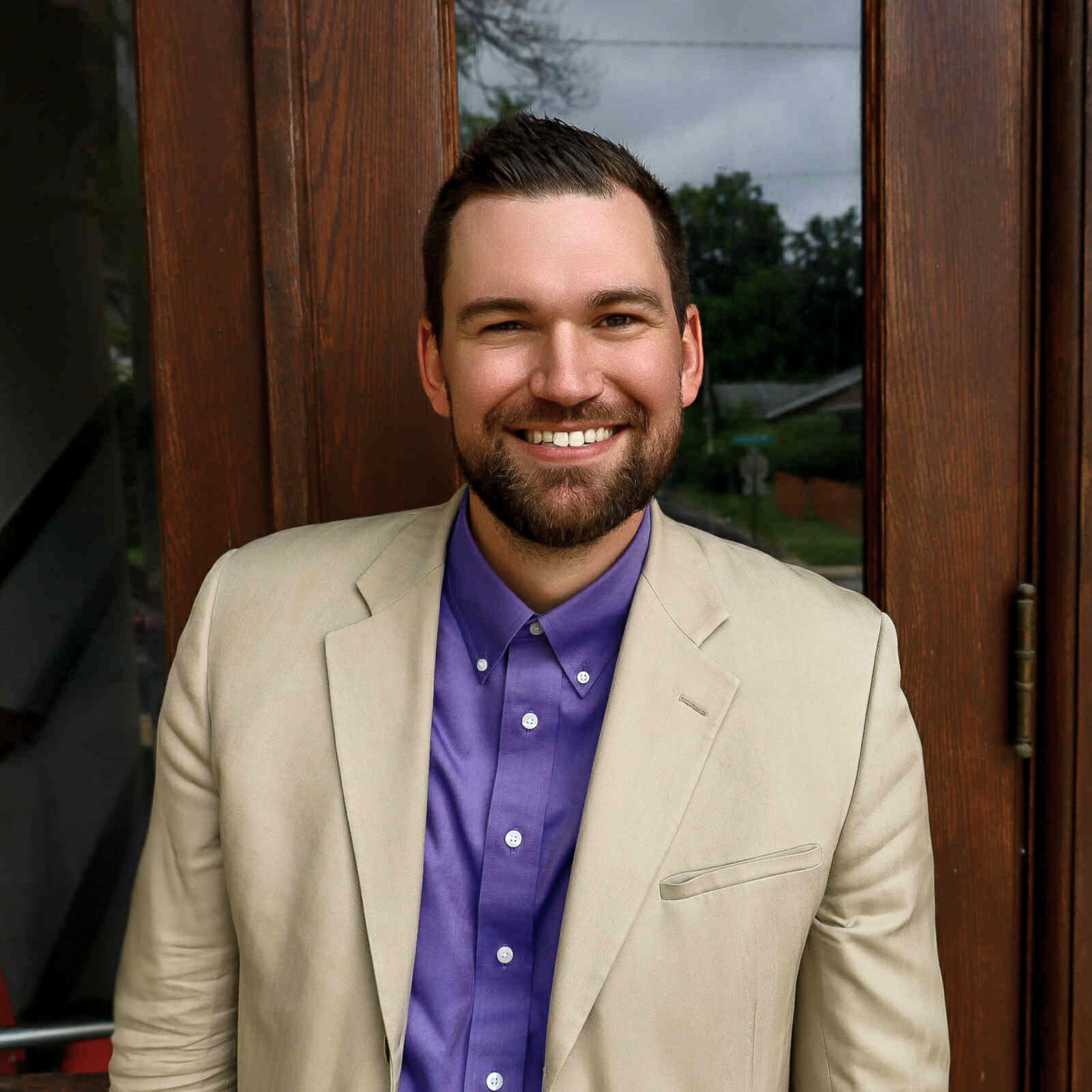 Close-up of a smiling man with short dark hair, beard, and mustache wearing a beige blazer and purple shirt, standing in front of a wooden window with trees in the background.