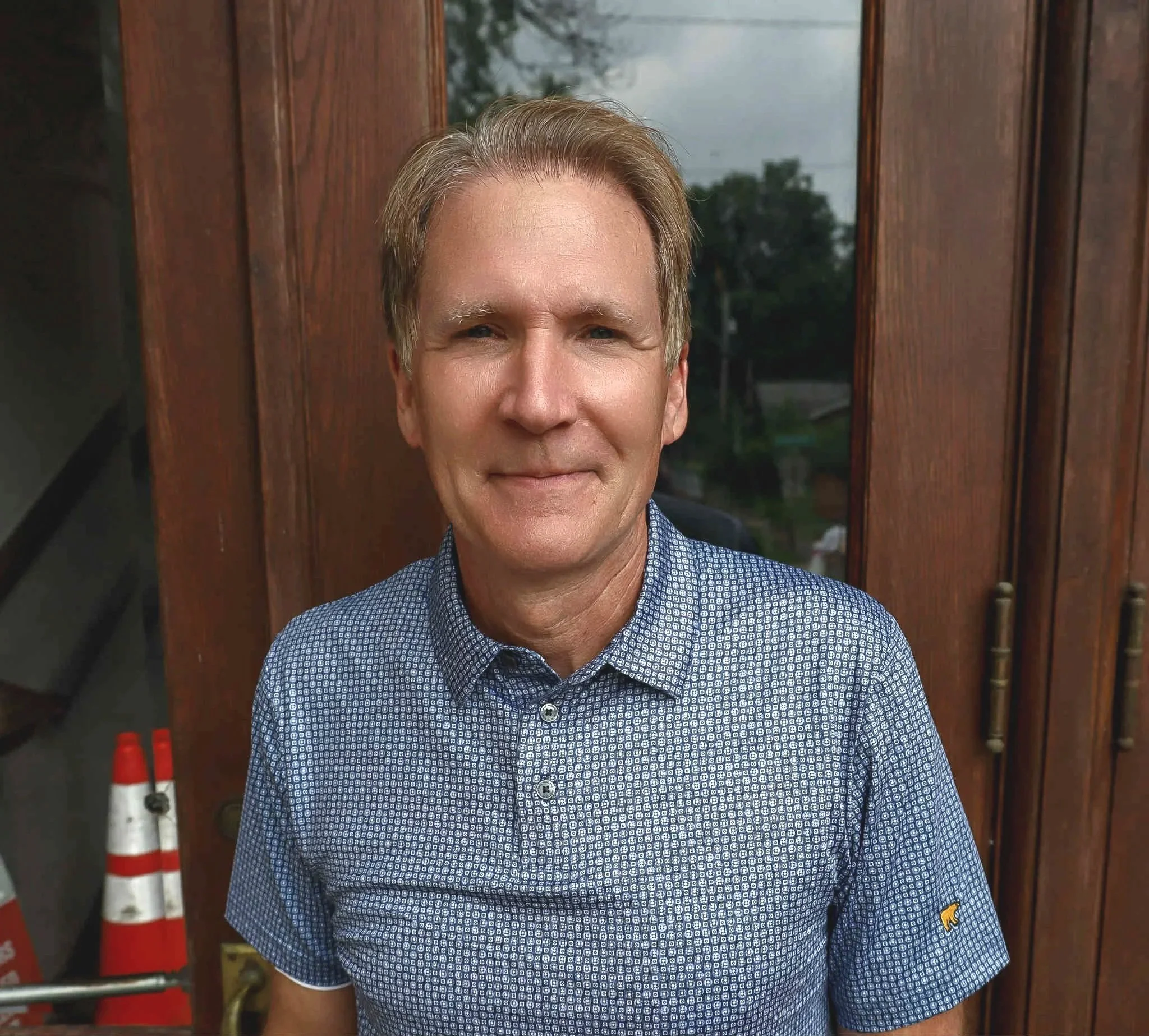 Close-up of a middle-aged man with light brown hair, wearing a blue patterned shirt, standing in front of a wooden door with a glass window. The man is smiling slightly and looking directly at the camera.