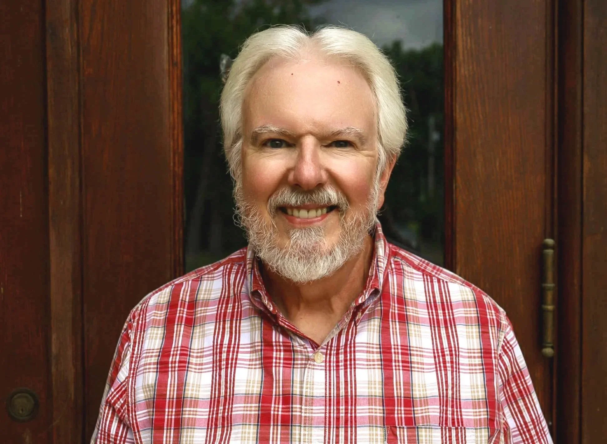A cheerful older man with gray hair, beard, and mustache, wearing a red and white plaid shirt, stands outside in front of a wooden door or wall.