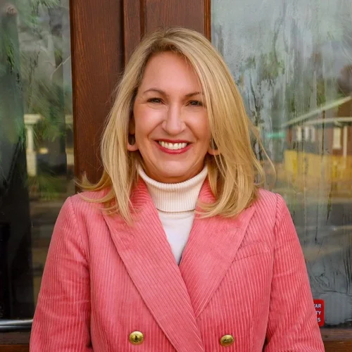 A woman with blonde hair smiling, wearing a pink blazer and white turtleneck, standing outside near a wooden door with a glass window.