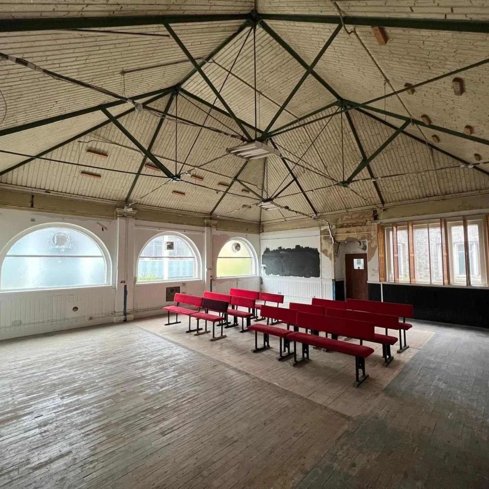 Empty room with wooden floors, arched windows, red seats arranged in rows, and an exposed ceiling with structural beams.