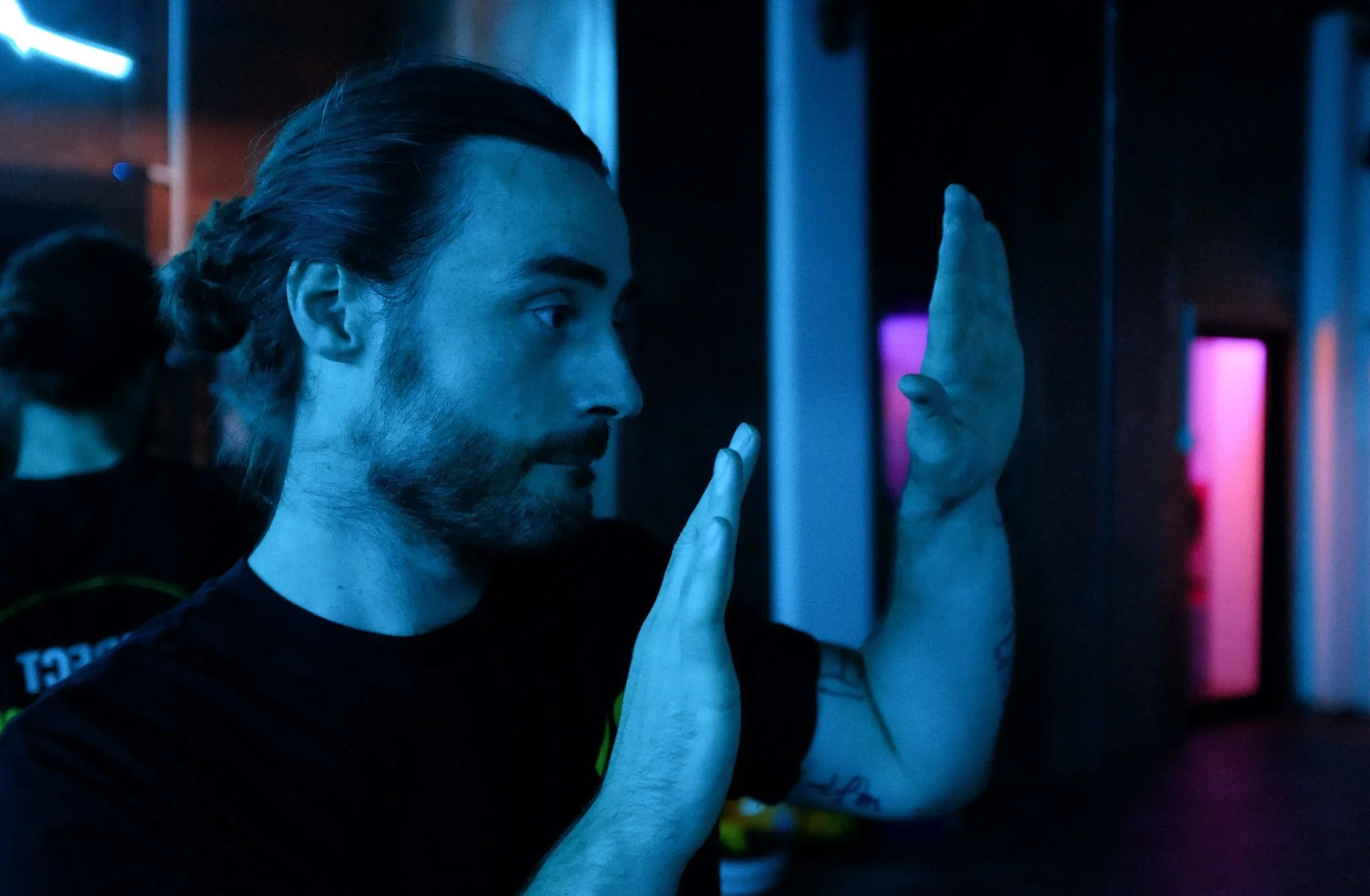 A man with long hair tied back in a bun, wearing a dark sports shirt, is in a dark room with neon lighting. He is facing sideways and has his hands raised in front of him, appearing to be practicing tai chi or a similar meditative movement.