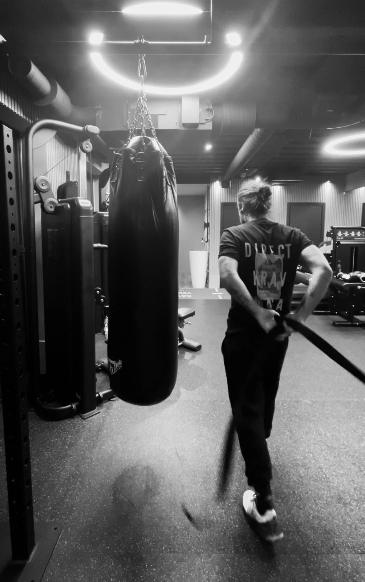 A person in a gym, wearing a shirt with the word 'DIRECT' on the back, is working out with a battle rope. A punching bag hangs nearby, and gym equipment is visible in the background. The environment has a modern, industrial style with circular ceiling lights.