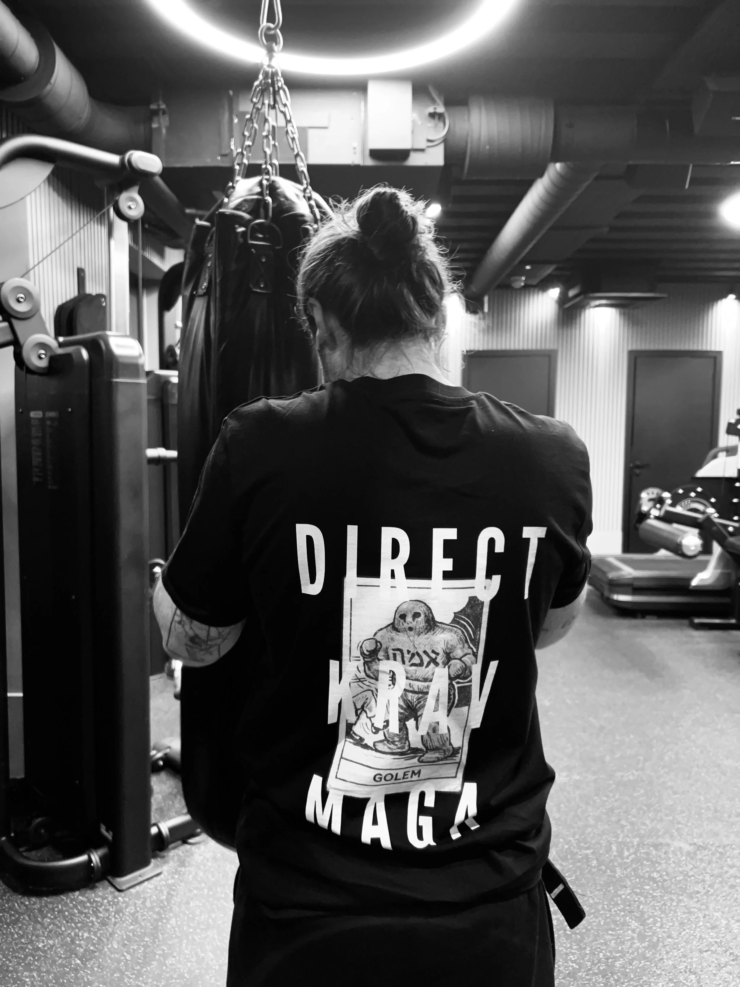 Back of a woman with hair in a bun, wearing a black T-shirt with an illustration of a boxer dog and the words 'DIRECT MAGA' on it, in a gym setting with exercise equipment and hanging punching bag.
