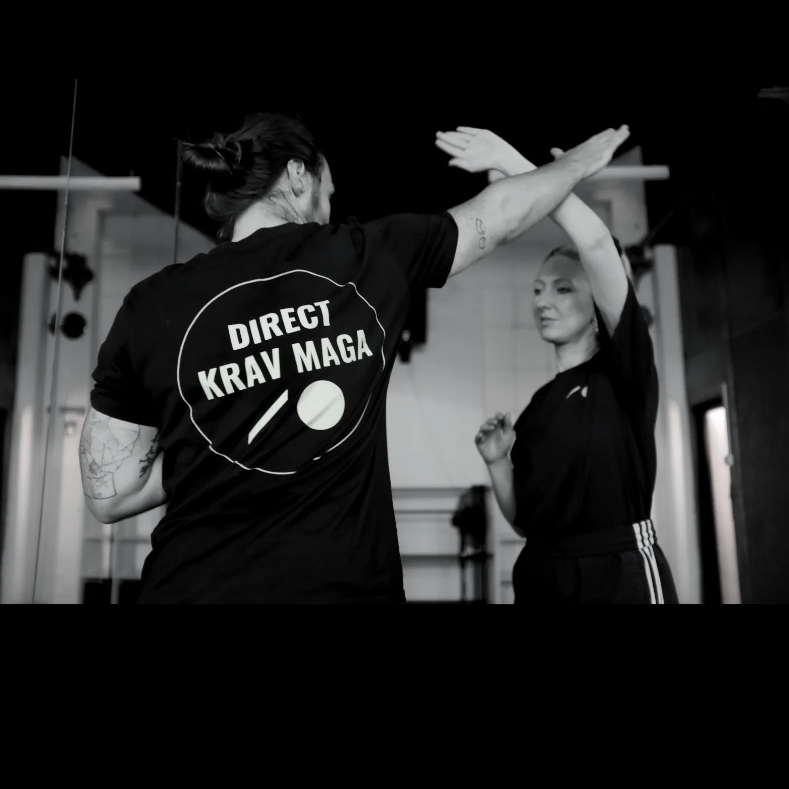 Two martial artists practicing Krav Maga in a gym, one giving a high five to the other, both wearing black shirts and pants.