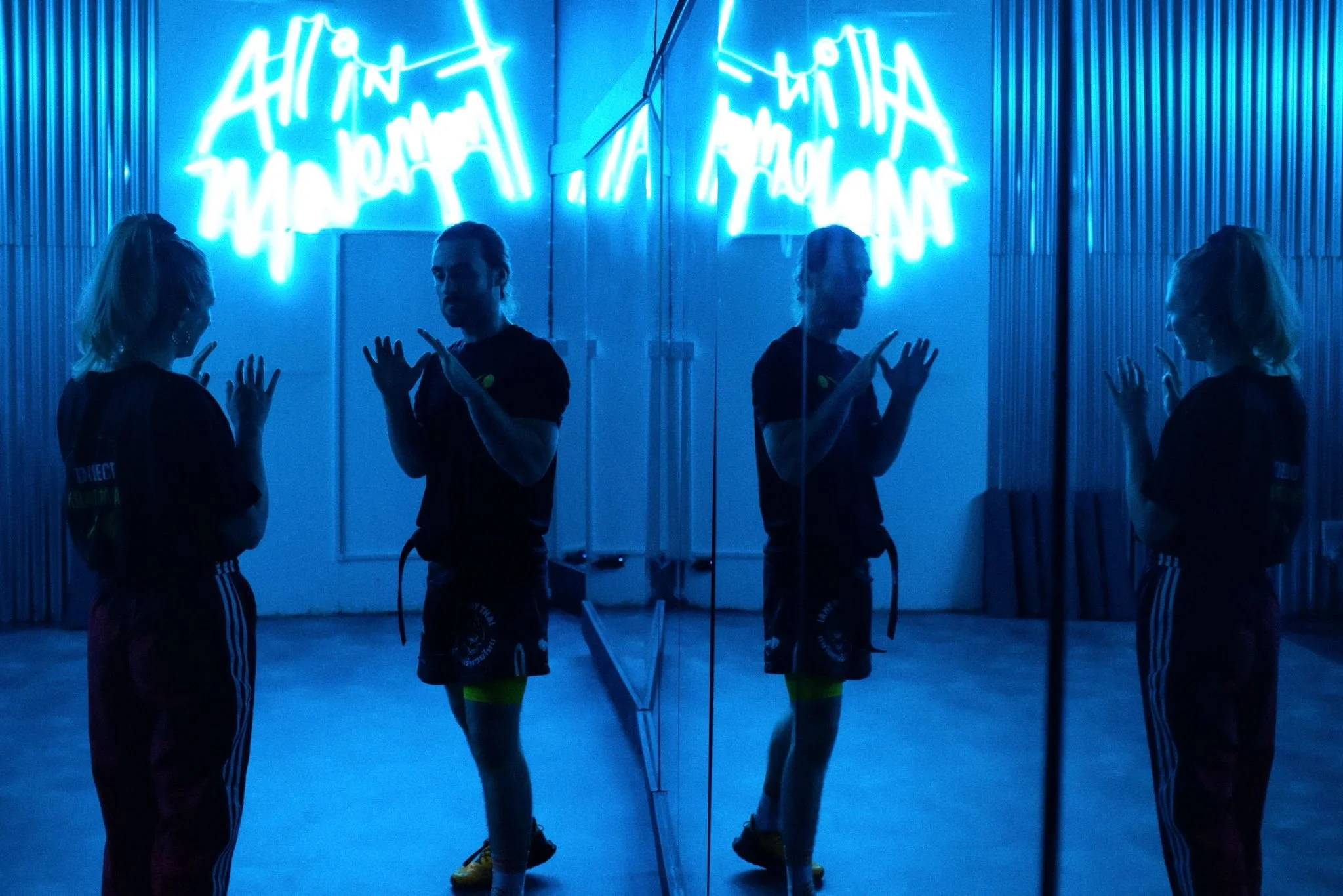 Two women in athletic clothing stand facing each other in front of a mirror with blue neon lights. Their reflections are visible, and they appear to be in a dance or fitness studio with a metallic wall background.