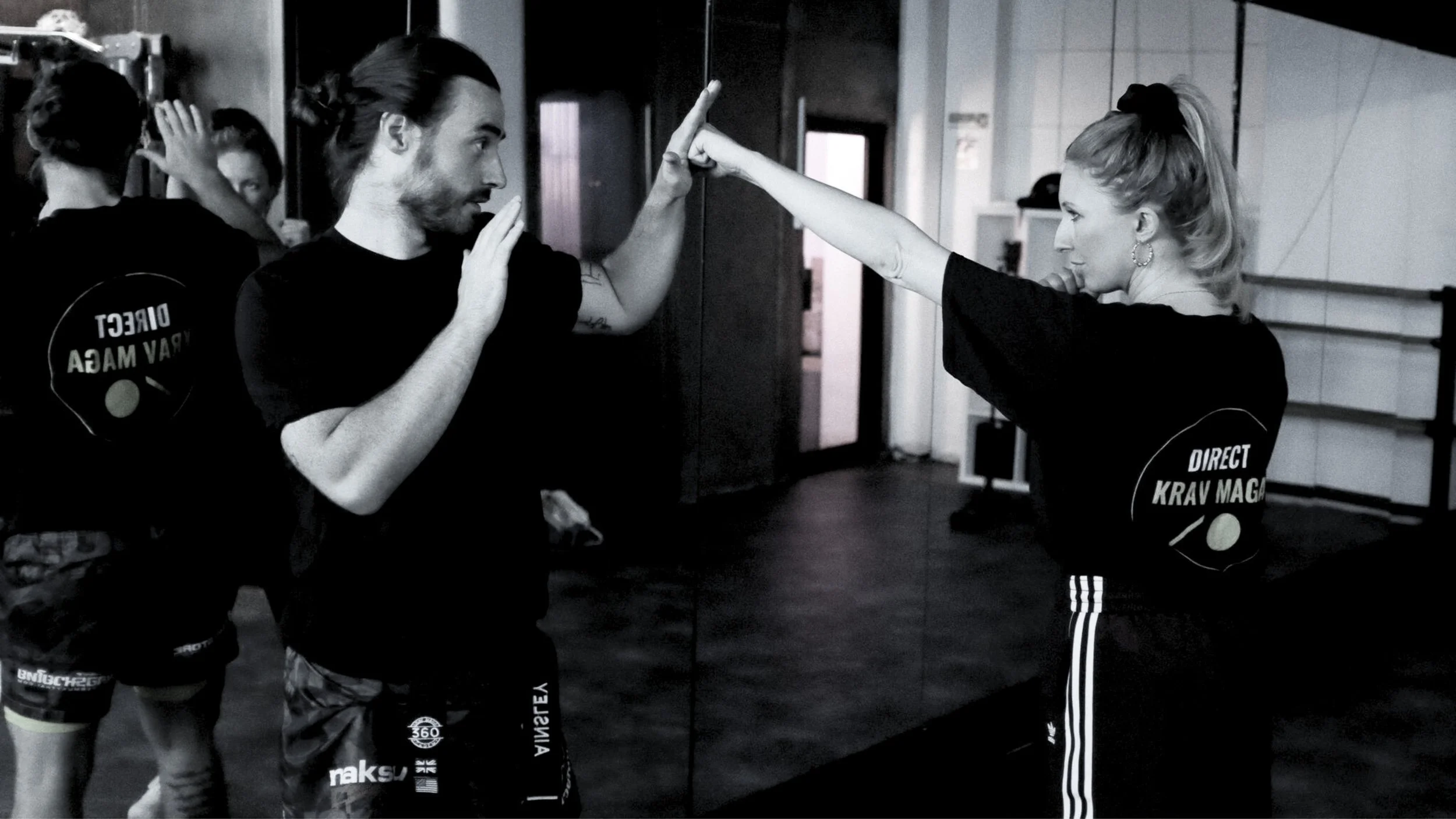 Two individuals practicing Krav Maga in front of a mirror, engaging in a defensive sparring move in a martial arts gym.