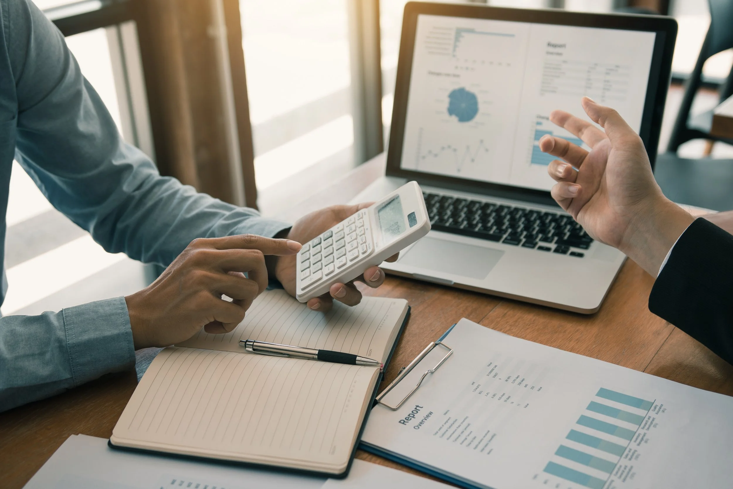 Two people analyzing financial reports with charts and graphs on a laptop, calculator, notebooks, and papers on a wooden desk.