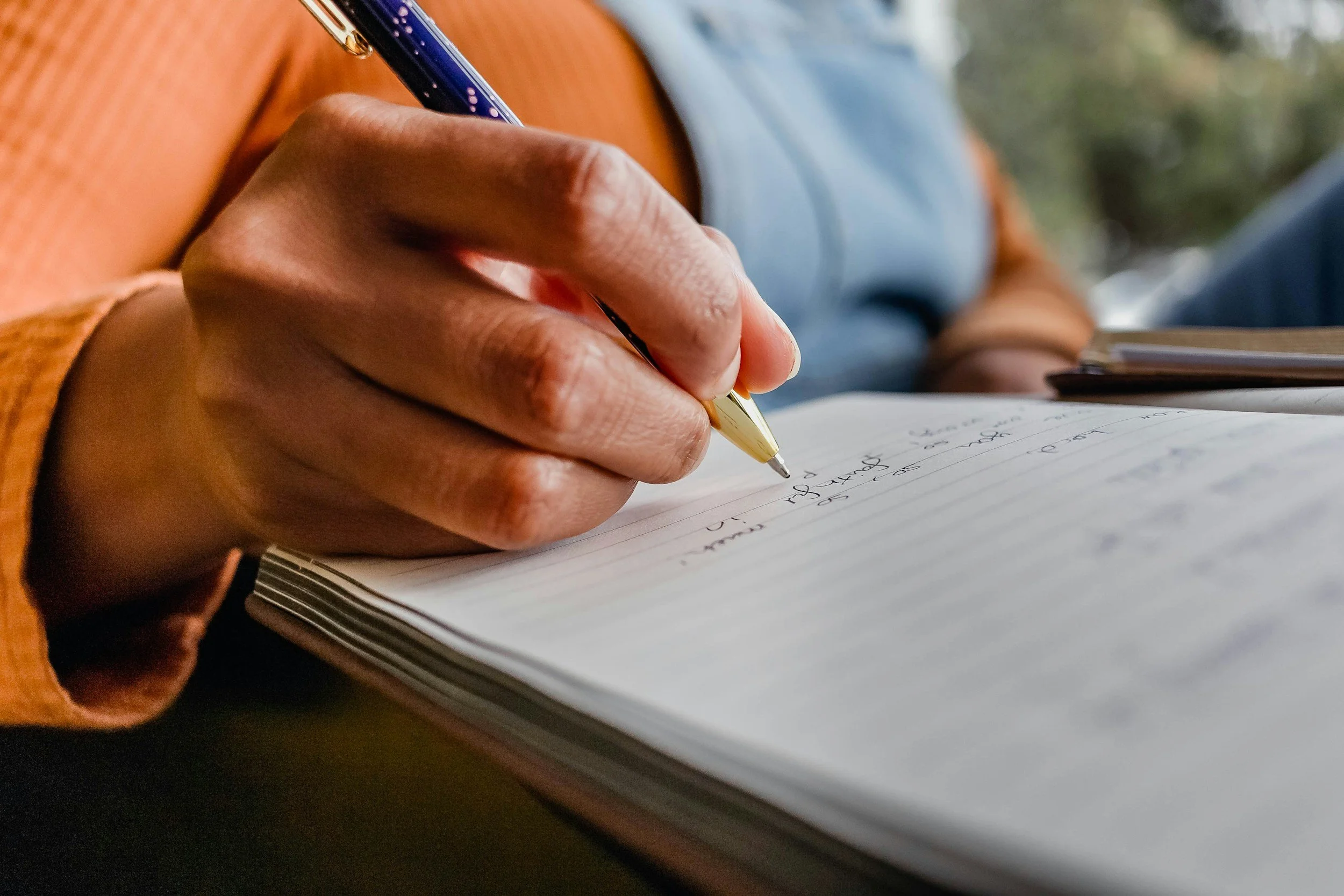 Close-up of a person's hand writing with a gold pen in a lined notebook, with an orange sleeve and a blue shirt visible.