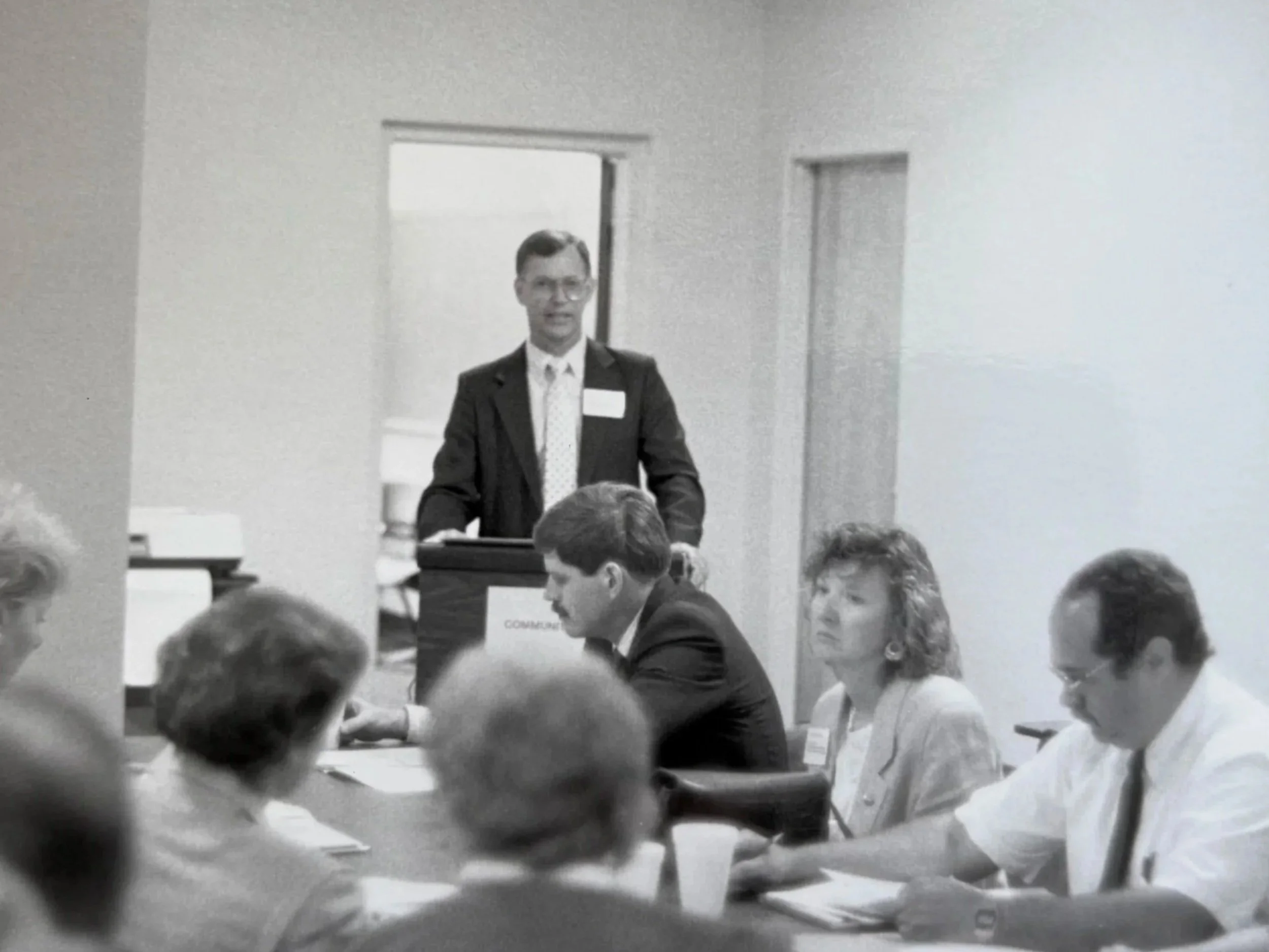 A man in a suit and glasses standing behind a podium, speaking to an audience at a conference or seminar.