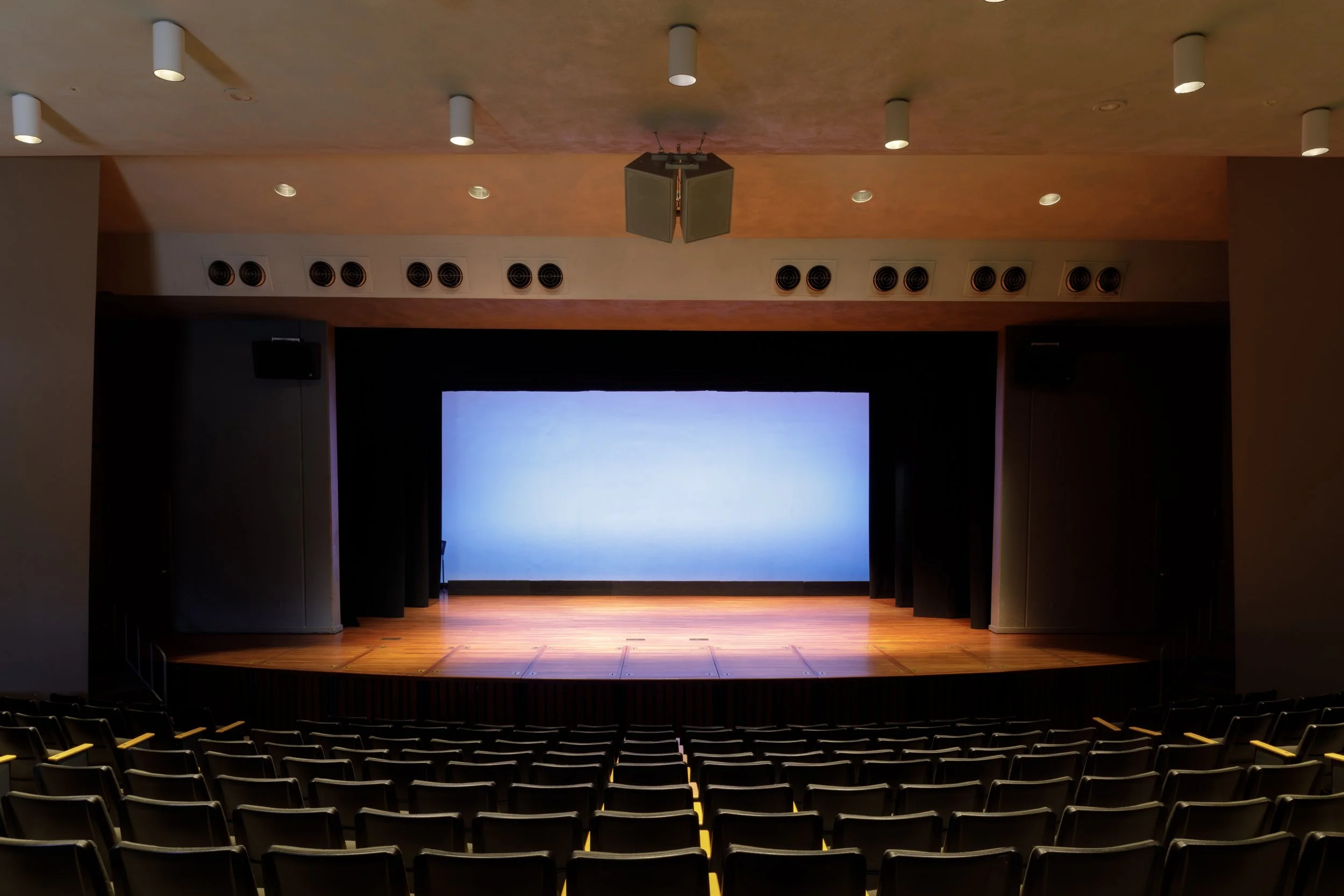An empty theater stage with a large screen behind it, surrounded by rows of theater seats. The ceiling has multiple stage lights and ventilation vents.