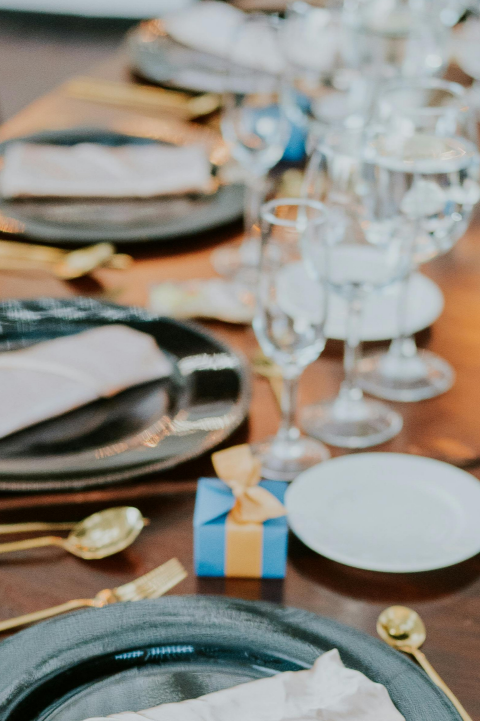 A festive table setting with black, gold, and white plates, gold utensils, clear glassware, a small blue gift box with a gold ribbon, and a white candle holder with lit candles.
