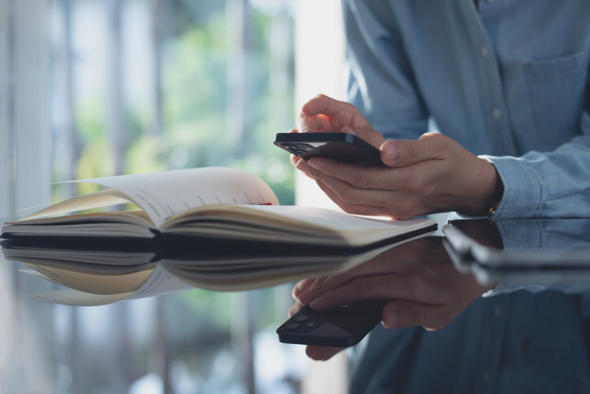 Person holding a smartphone above an open notebook with writing, sitting at a desk near a window with blurred greenery outside.
