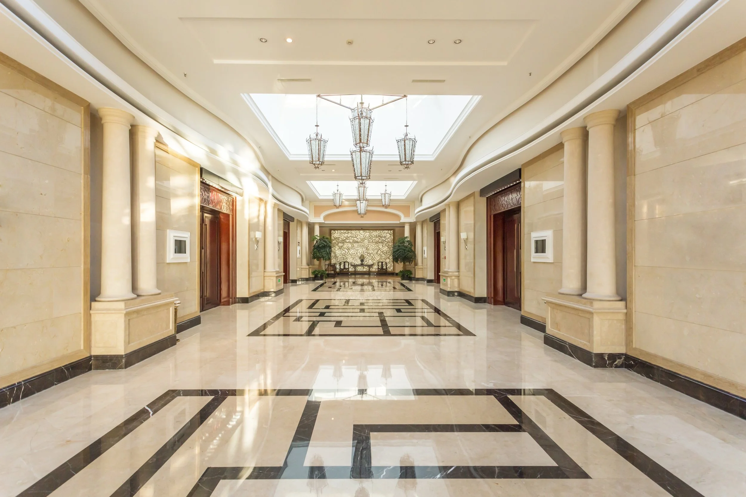 A luxurious hotel lobby with beige marble walls and floors, black marble accents, tall columns, ornate pendant lighting, and greenery in the background.