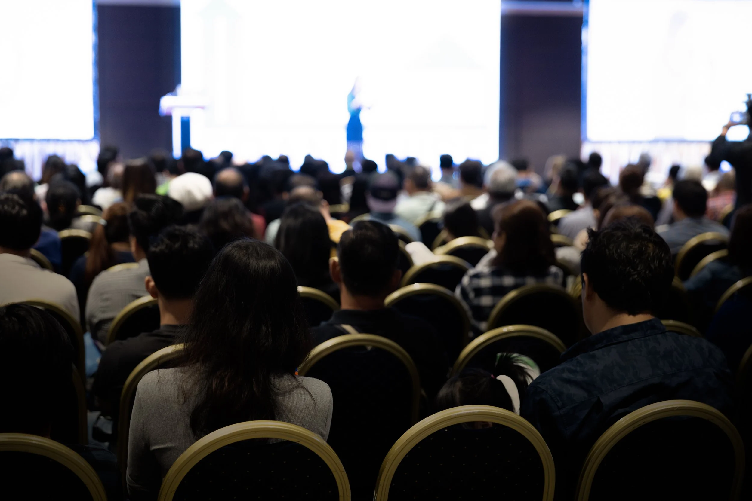 A large audience seated in a conference hall, facing a stage with bright screens and a person presenting.