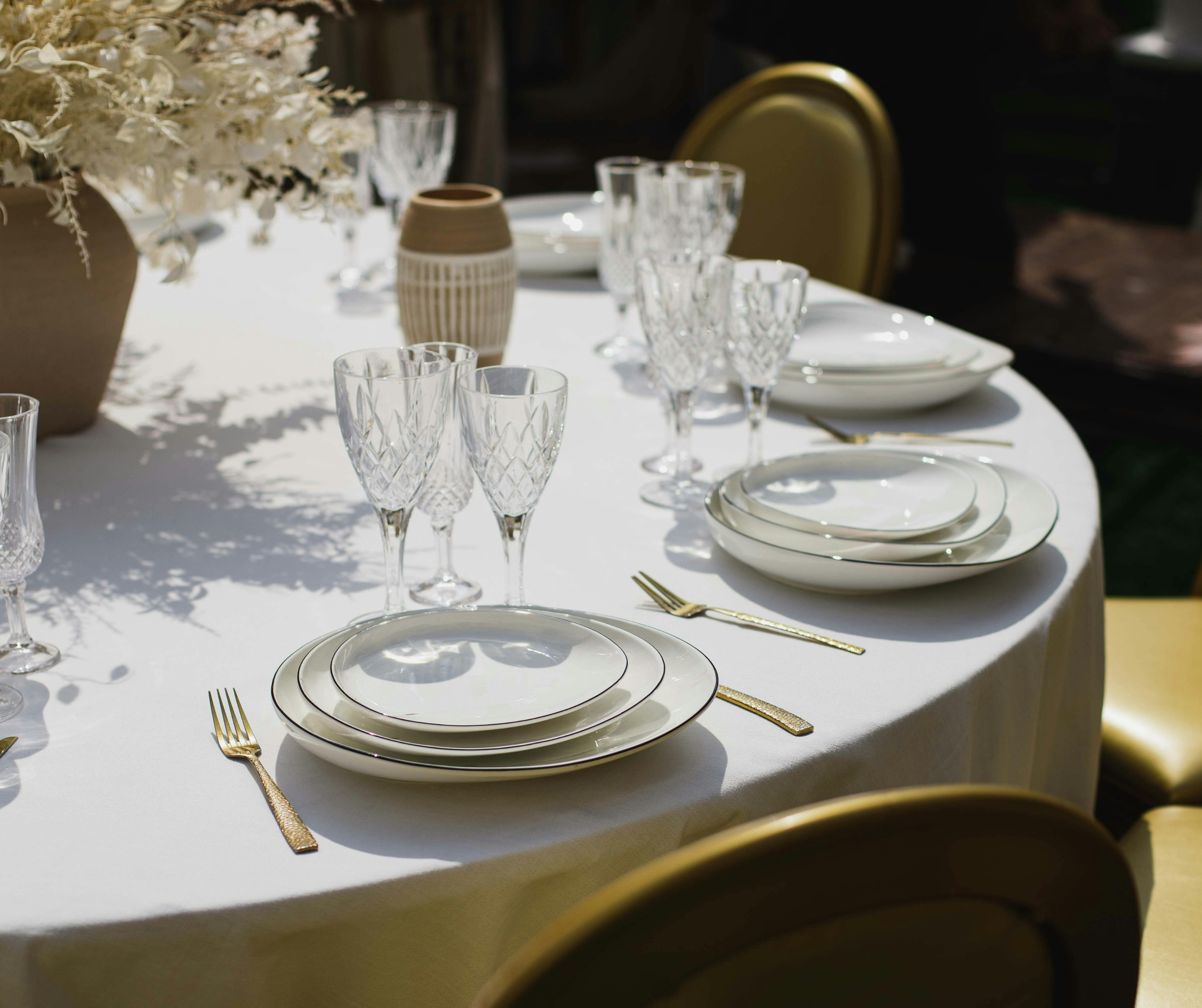 Elegant table setting with white plates, gold utensils, clear crystal glasses, and a beige vase with dried flowers on a white tablecloth.