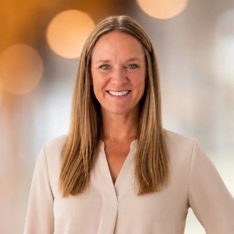A smiling woman with long light brown hair standing in front of a blurred background with warm circular lights.