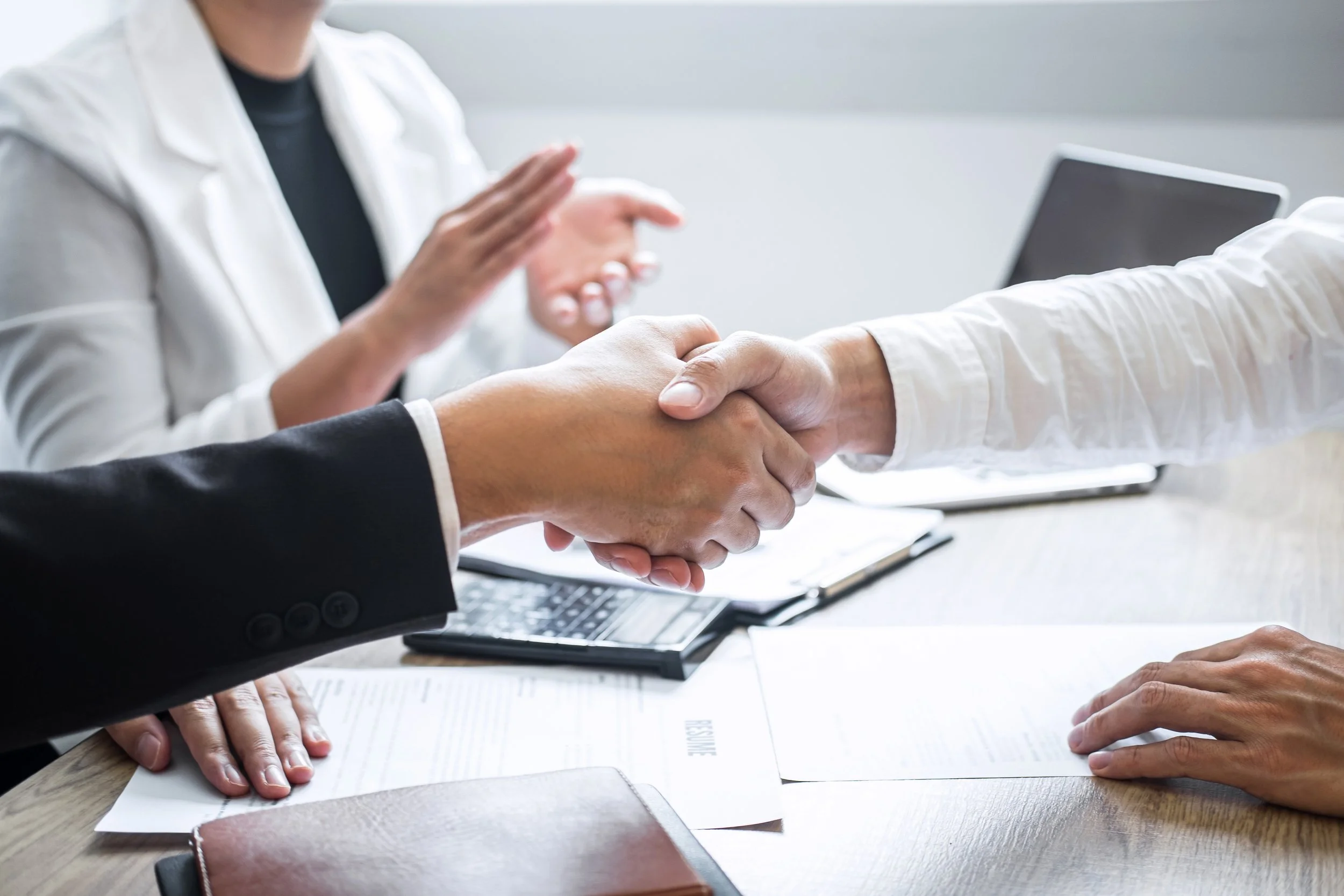 Two people shaking hands over a meeting table, with documents, a laptop, and a notebook present, suggesting a business meeting or agreement.