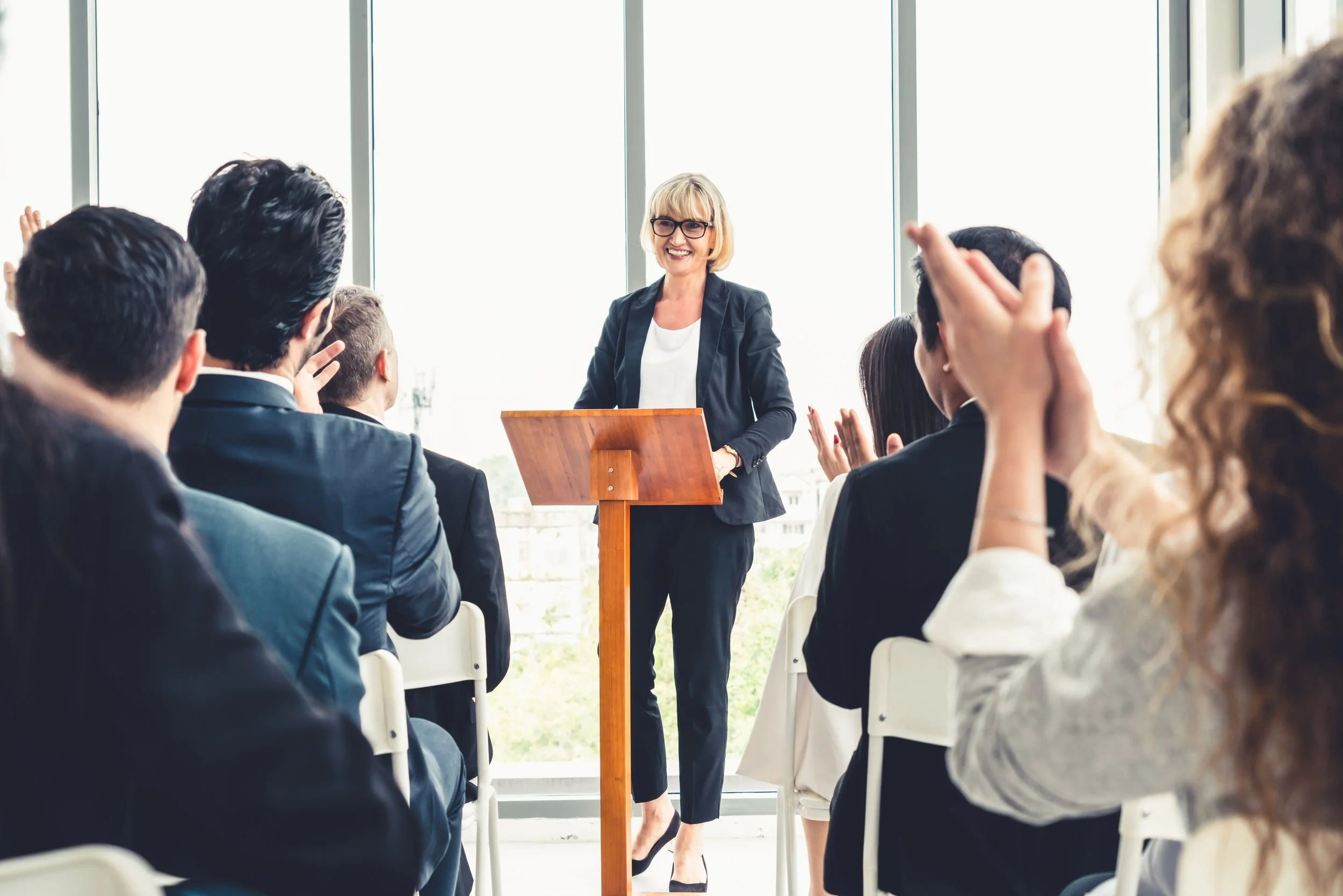 A woman in a black blazer and glasses standing at a wooden podium, smiling, in front of a large window with people seated and raising their hands in a meeting or conference setting.