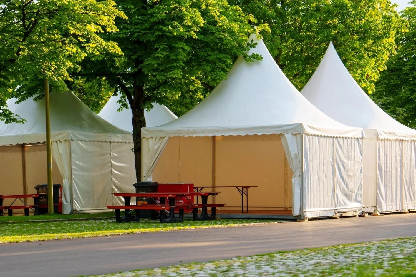 Several large white event tents set up outdoors among green trees, with red picnic tables and benches nearby.