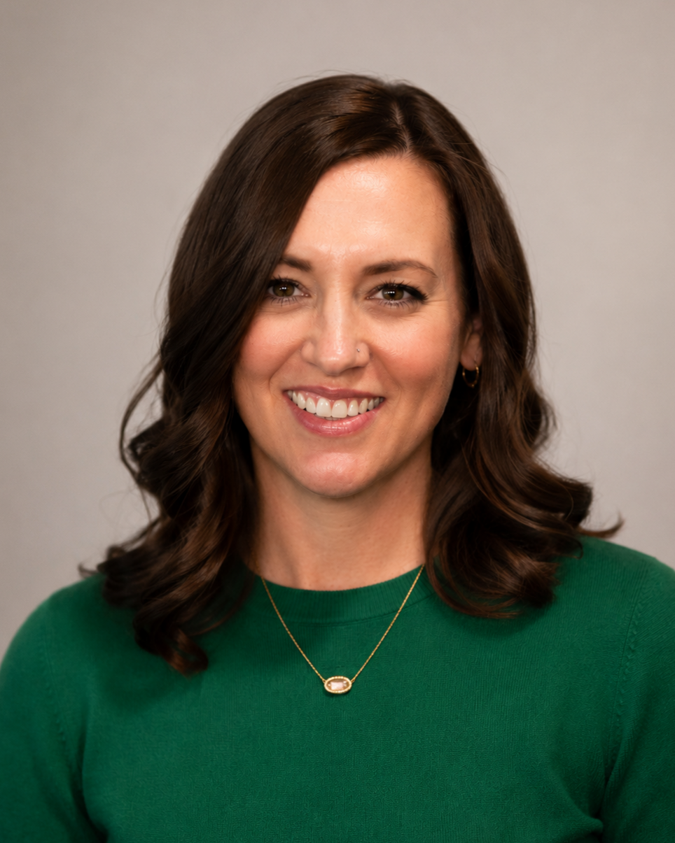 A woman with shoulder-length brown hair, wearing a green top and gold jewelry, smiling at the camera against a neutral background.