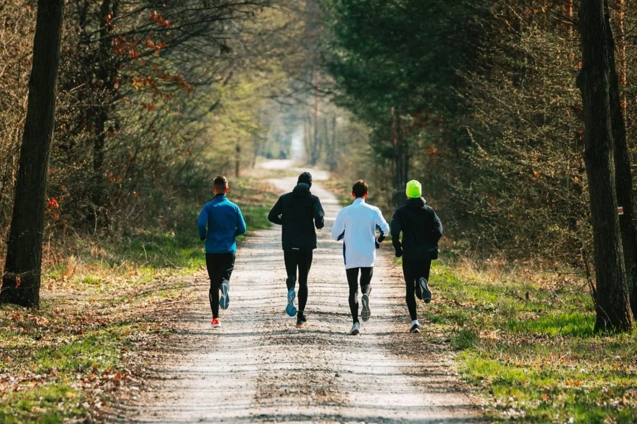 Four people jogging on a dirt trail through a wooded area with autumn trees.