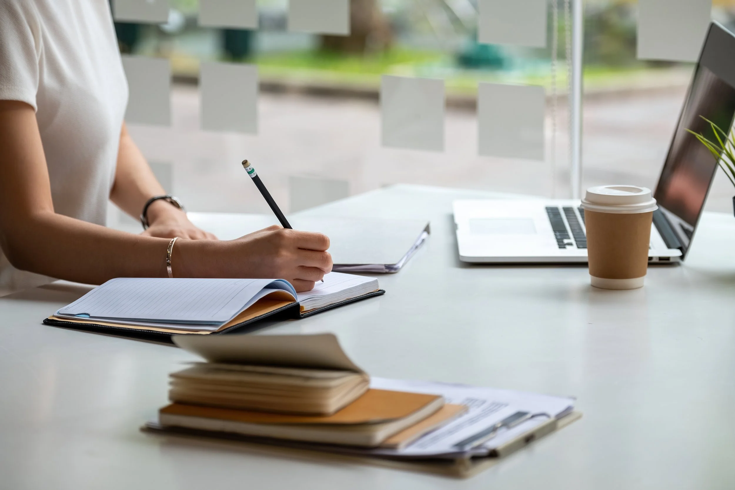 Person writing in a notebook at a desk with a laptop, disposable coffee cup, and stacks of paper and books, near a window with blurred outdoor scenery.