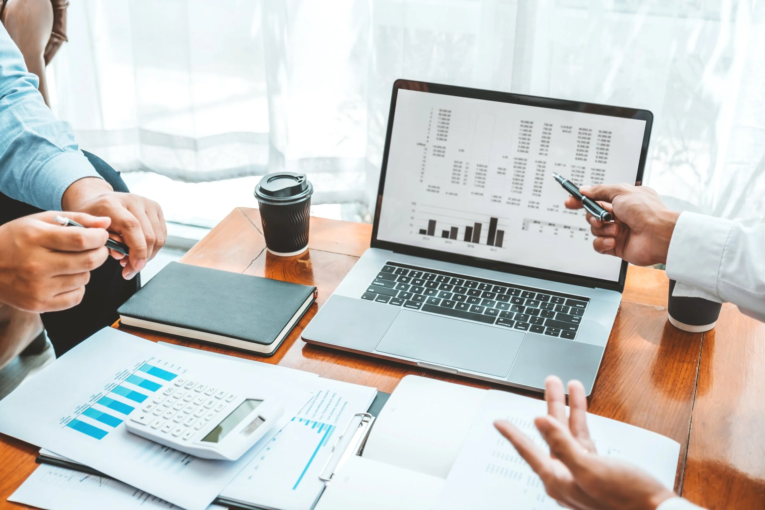 Business meeting with financial documents, graphs, calculator, and a laptop displaying spreadsheets, with people discussing and pointing at the screen.