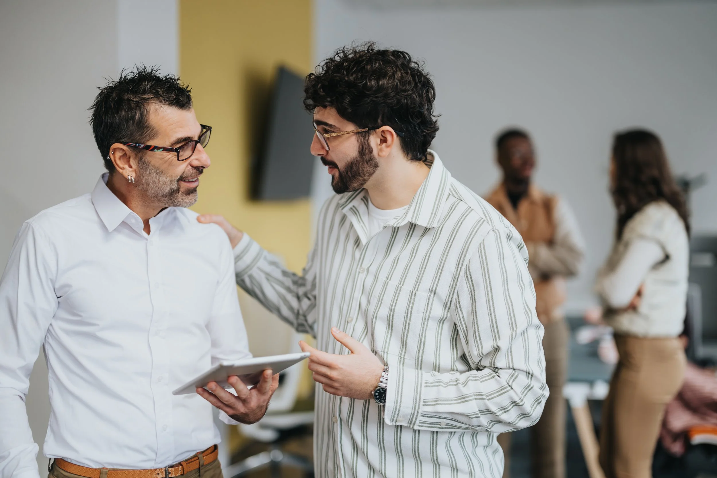 Two men are talking in a workplace setting, with one wearing a white shirt and glasses, and the other in a striped shirt and glasses, holding a tablet, while three other people are in the background.